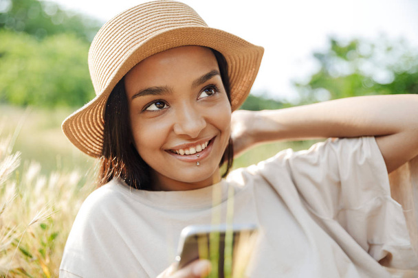Πορτρέτο της χαμογελαστή γυναίκα φορώντας ψάθινο καπέλο χρησιμοποιώντας το κινητό τηλέφωνο, ενώ βρίσκεται σε γρασίδι σε πράσινο πάρκο - Φωτογραφία, εικόνα