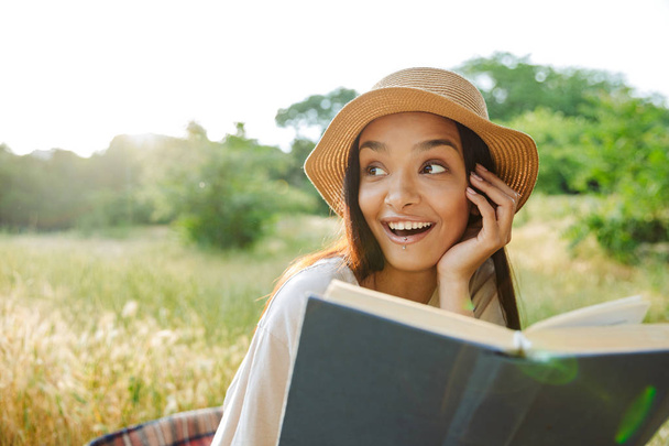 Портрет восторженной женщины в соломенной шляпе, читающей книгу, сидящей на траве в зеленом парке
 - Фото, изображение