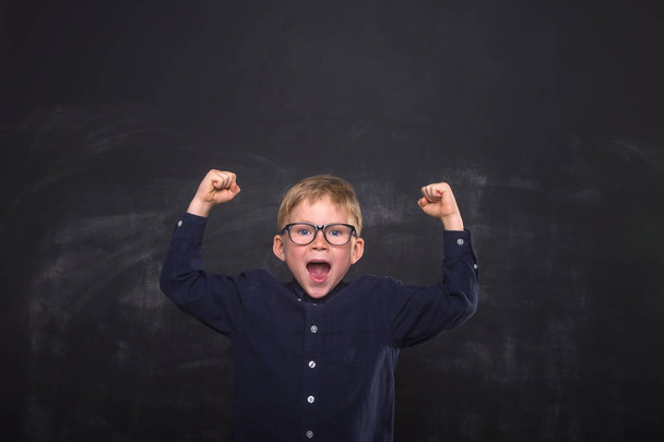 Happy school child boy in uniform and glasses against blackboard. Yeah! Strong kid raising hands up. Successful education. Stop bulling. - 写真・画像