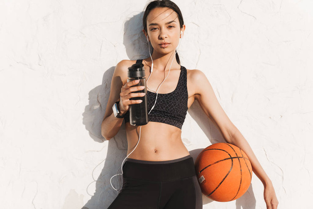 Amazing concentrated strong sports woman outdoors over white wall background posing listening music with earphones holding ball and bottle with water. - Foto, Bild