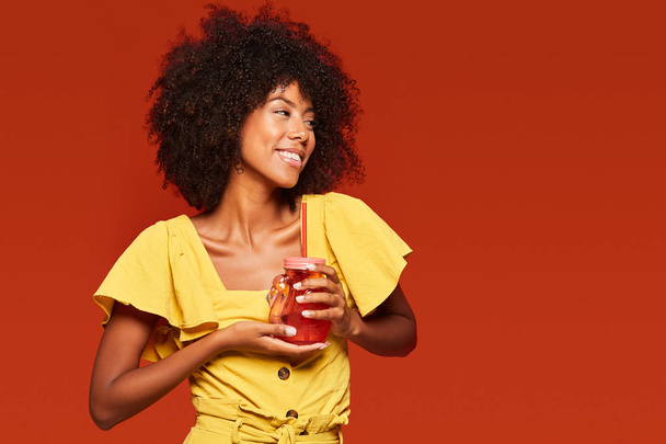 Querida mujer afroamericana con vello curado con jarro rojo con paja y disfrutando de una bebida en fondo rojo. - Foto, Imagen