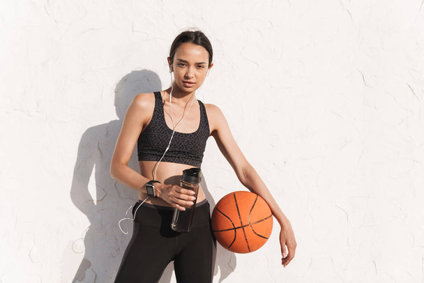 Amazing concentrated strong sports woman outdoors over white wall background posing listening music with earphones holding ball and bottle with water. - Foto, afbeelding