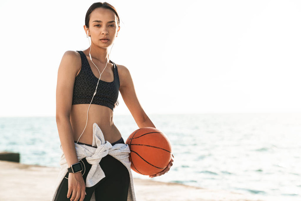 Beautiful strong sports woman outdoors at beach posing listening music with earphones holding basketball. - Φωτογραφία, εικόνα