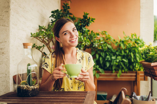 Romantic portrait of beautiful young woman wearing yellow dress, relaxing on the balcony between many green plants, holding cup of tea or coffee - Foto, Bild