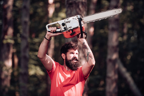 Stylish young man posing like lumberjack. Lumberjack on serious face carries chainsaw. Lumberjack worker standing in the forest with chainsaw. Man doing mans job. - Фото, изображение