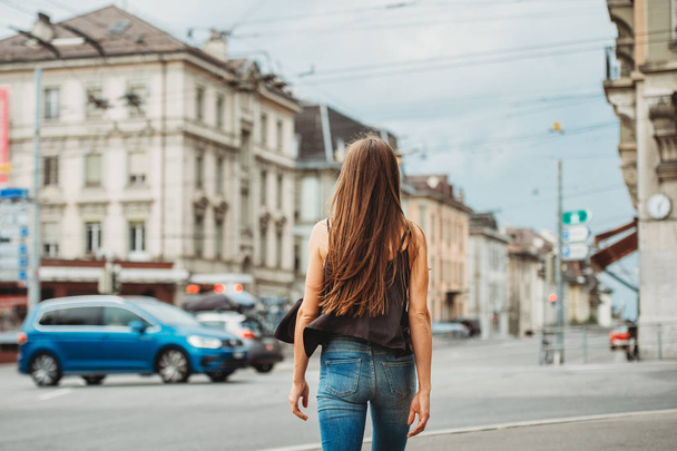 Outdoor portrait of beautiful young woman walking down the road in the city, wearing black cami top and denim jeans. Back view. Image taken in Lausanne downtown, place Bel Air, Switzerland - Фото, зображення