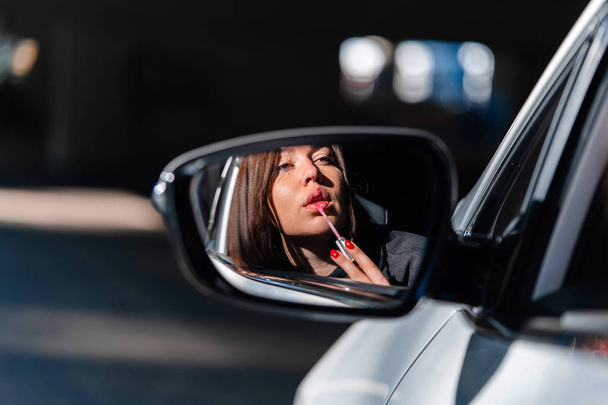 Confident and beautiful young business woman painting her lips in new car and looking into Side view mirror. - Photo, image