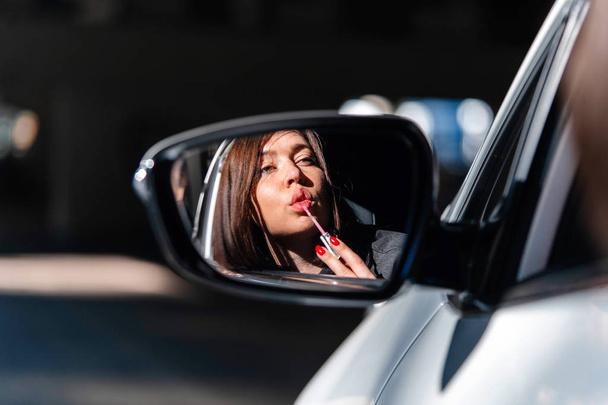 Confident and beautiful young business woman painting her lips in new car and looking into Side view mirror. - Photo, image