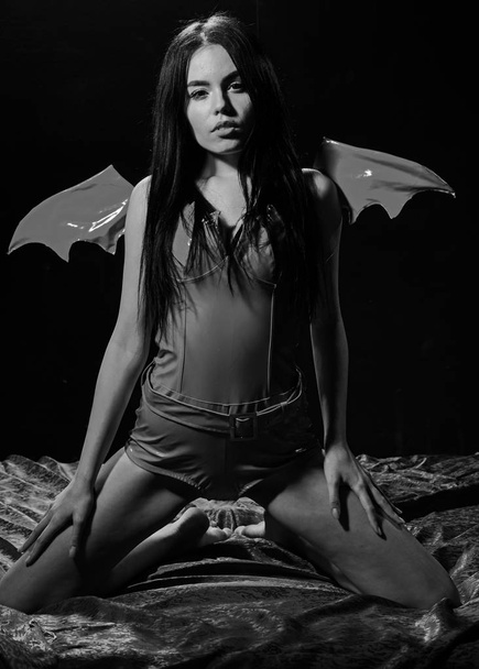 Lady sexi dressed as demon, devil, black background. Sexy devil concept. Demon sexy girl sit on silk sheets. Woman on passionate face play role game. Girl sexy demon with wings, devil full of desire - Foto, afbeelding