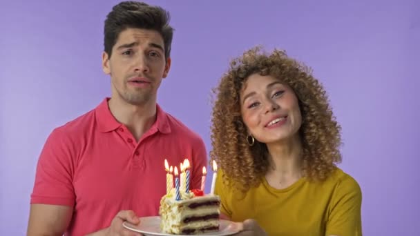 Happy young lovely couple giving a birthday cake and singing happy birthday while looking at the camera over purple background isolated - Footage, Video