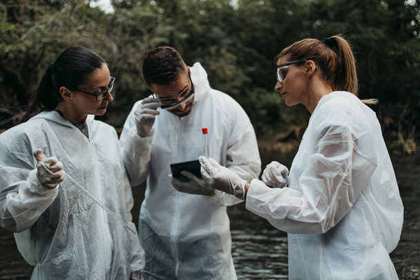 Scientists biologists and researchers in protective suits taking water samples from polluted river. - Фото, изображение