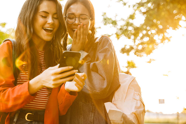 Image of two delighted girls laughing and typing on cellphone while walking in green park - 写真・画像