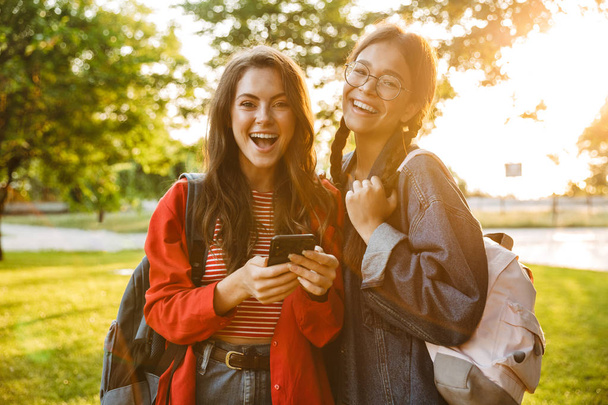 Image of two delighted girls smiling and typing on cellphone while walking in green park - Foto, Imagem