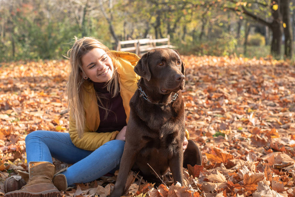 A beautiful young blond-haired woman is having fun with her big brown labrador dog in the park - Photo, Image