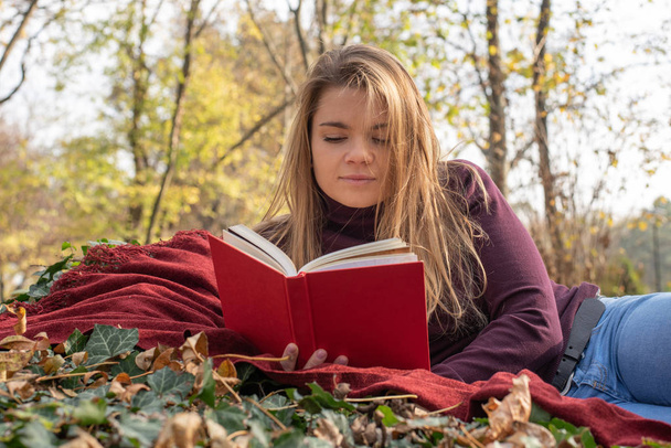 A beautiful young blond-haired woman is reading in the autumn park - Photo, Image