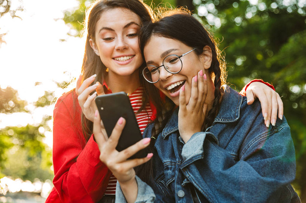Image of two excited girls students using cellphone and hugging while sitting on railing in green park - Fotografie, Obrázek