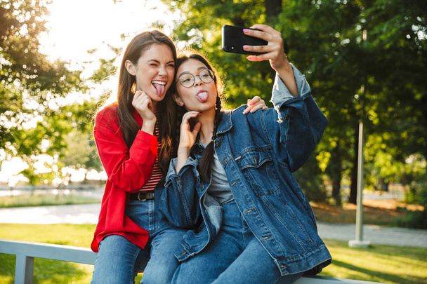 Image of two funny girls taking selfie photo on cellphone with tongues sticking out while sitting on railing in green park - Foto, afbeelding