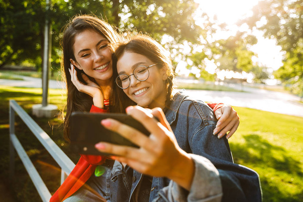 Image of two pretty girls taking selfie photo on cellphone and smiling while sitting on railing in green park - Fotoğraf, Görsel