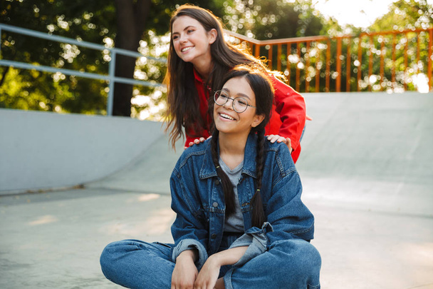 Image of two pretty girls laughing and riding skateboard together in skate park - Foto, afbeelding