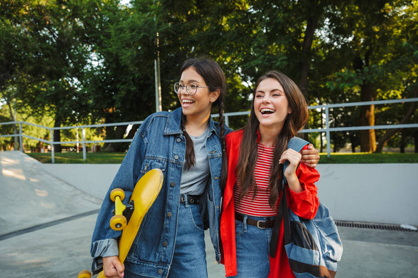 Image of two adorable girls smiling and hugging together while holding skateboard in skate park - Foto, afbeelding