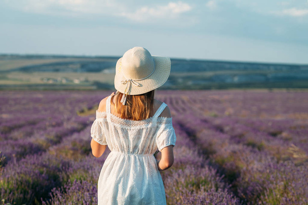 A young girl with a hat and a white dress stands with her back in a lavender field - Фото, зображення