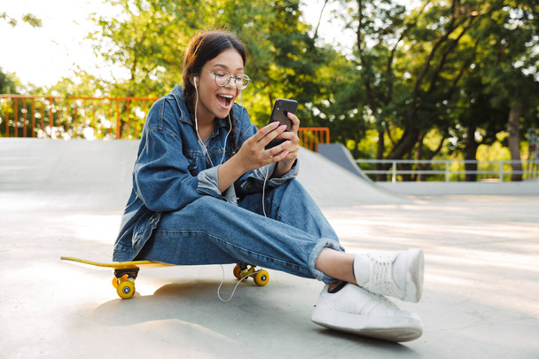 Image of excited girl using smartphone and earphones while sitting on skateboard in skate park - Fotó, kép
