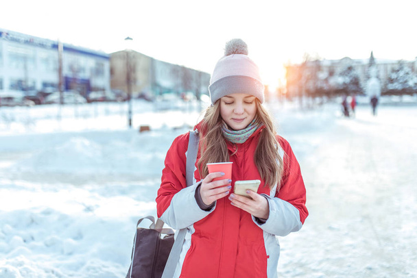 girl winter city background snow snowdrifts, stands warm jacket hat, holds cup tea with hot coffee drinks, writes message smartphone, social networks read online watch video on phone. - Photo, Image