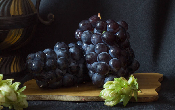 Dense small grapes in droplets of water on a wooden board and a little hops on a black background. - Foto, Bild