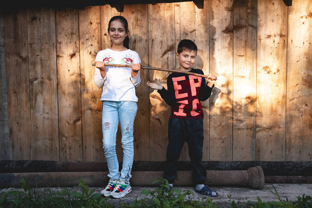 Schattige kleine jongen en mooi meisje in Casual kleding houden in handen houten stok, op zoek met een glimlach op de camera, staande op de houten achtergrond buiten - Foto, afbeelding