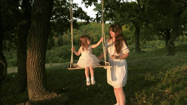 ママは太陽の下で木の下でブランコで彼女の娘を振る。クローズ アップ。母と赤ちゃんは森のオークの枝にロープスイングに乗ります。女の子は笑い、喜ぶ。自然の中で、公園で家族の楽しみ。暖かい夏の日. - 写真・画像