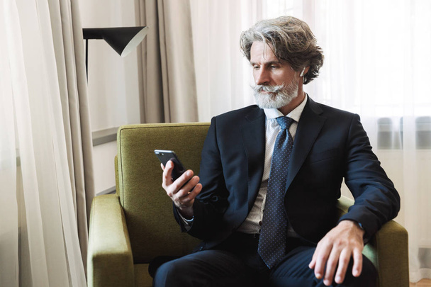 Handsome bearded mature grey-haired businessman posing indoors at home near window dressed in formal clothes using mobile phone. - Foto, Bild