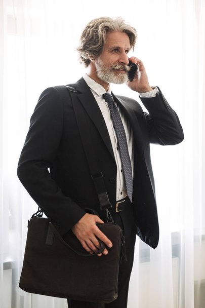 Serious handsome grey-haired senior businessman indoors at home dressed in formal clothes talking by mobile phone holding bag. - Fotografie, Obrázek