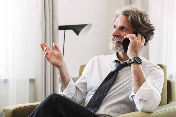 Image of joyful mature man talking on cellphone while sitting on - Fotó, kép