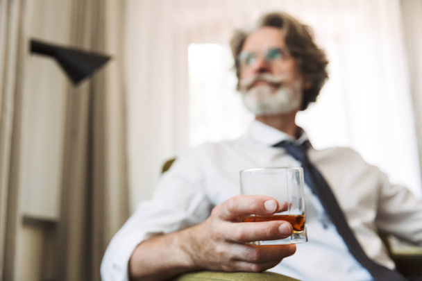 Handsome senior gray-haired business man sitting in chair indoors at home drinking alcohol. - Fotoğraf, Görsel
