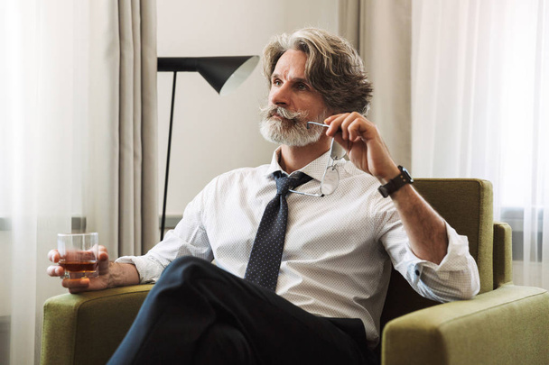 Handsome senior gray-haired business man sitting in chair indoors at home drinking alcohol. - 写真・画像
