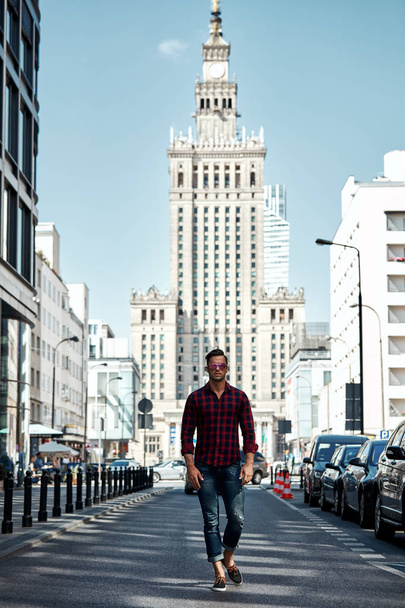 Joven hombre guapo en camisa a cuadros y jeans posando sobre fondo urbano
 - Foto, Imagen