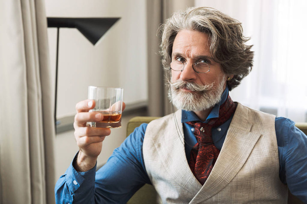 Portrait of handsome adult businessman drinking alcohol while sitting on armchair in hotel apartment - Φωτογραφία, εικόνα