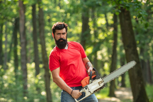 A handsome young man with a beard carries a tree. Illegal logging continues today. Lumberjack in the woods with chainsaw axe. - Fotografie, Obrázek