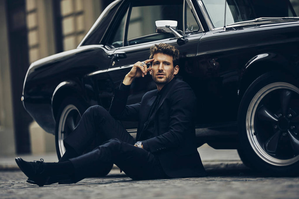 Handsome man in black suit sitting on street near old classic car - Foto, imagen