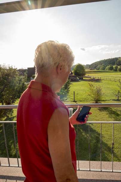 Seniorin mit weißem Haar steht auf dem Balkon und schaut auf ihr Handy. - Foto, Bild
