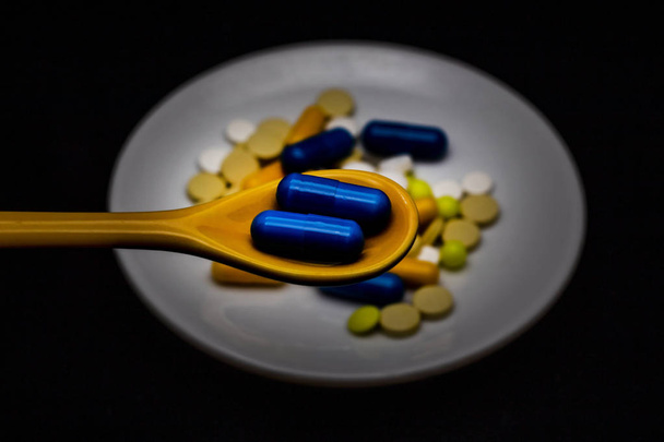 Spoon kapszulák, mögötte egy tányér, különböző színű gyógyszeripari tabletták és kapszulák. Sötét fény, fekete háttér. - Fotó, kép