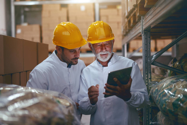 Two hardworking warehouse workers standing in warehouse and checking inventory. Older one holding tablet while younger one looking at it. - Photo, Image