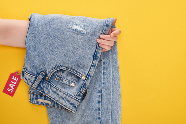 cropped view of woman holding jeans with sale label on yellow background - Photo, Image