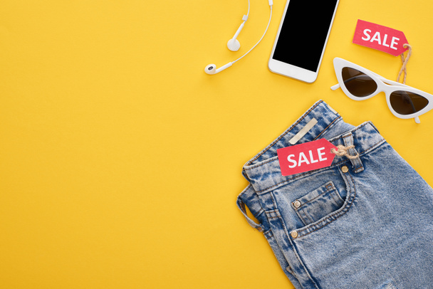 Ansicht von Jeans, Brille und Smartphone mit Kopfhörern mit Verkaufsetiketten auf gelbem Hintergrund - Foto, Bild