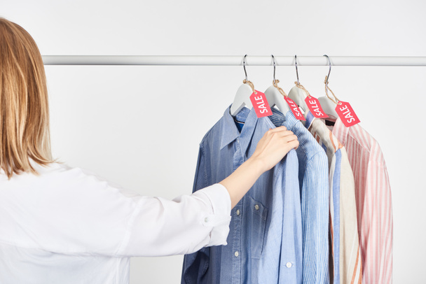 cropped view of blonde woman near elegant shirts hanging with sale labels isolated on white - Photo, Image