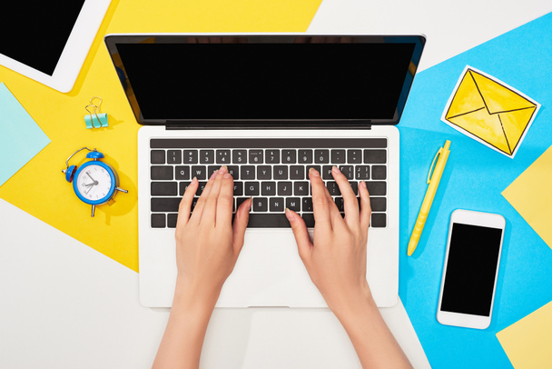 cropped view of woman using laptop near office supplies and mail icon on yellow, blue and white background - Fotó, kép