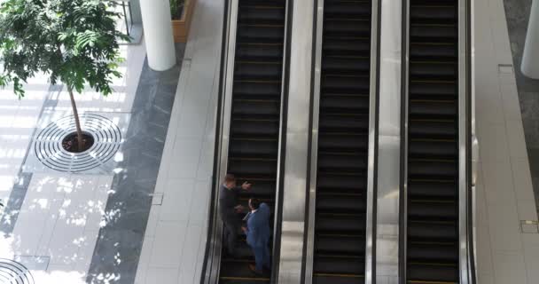 High angle view of a young Caucasian businessman and a middle aged Caucasian businessman talking as they stand going up an escalator in a modern office building, one holding a laptop and one holding a tablet computer - Filmati, video