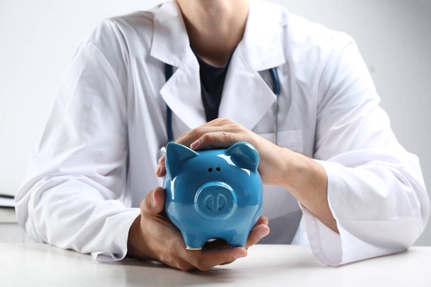 Doctor with piggybank at white table indoors, closeup. Medical insurance concept - 写真・画像