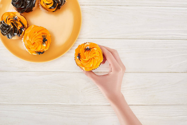cropped view of woman taking delicious cupcake with spiders from orange plate on white wooden table, Halloween treat - Photo, Image