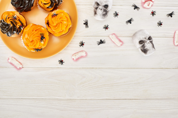 верхний вид на кексы на оранжевой тарелке, желейные зубы, черепа и пауки на белом деревянном столе, Хэллоуин удовольствие
 - Фото, изображение
